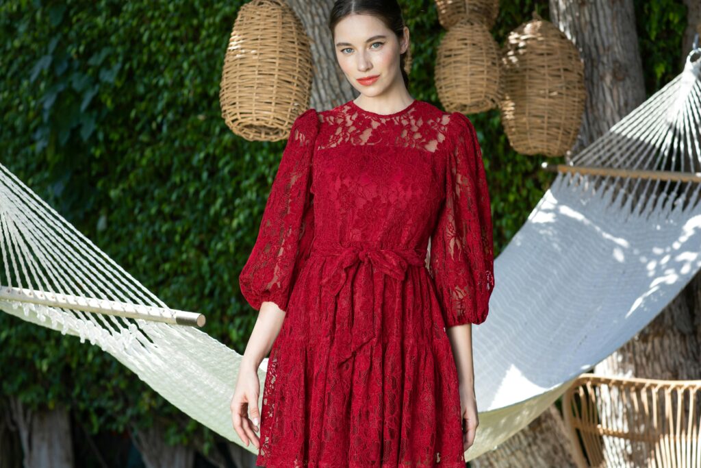 Stylish woman in red lace dress standing outdoors with wicker baskets and hammock.