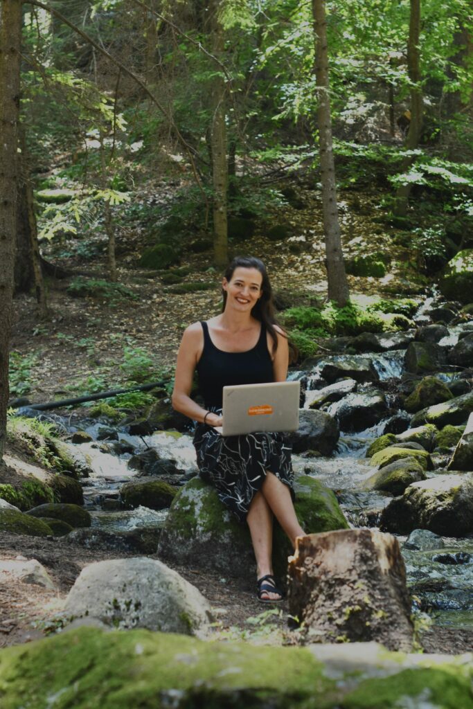 A woman enjoying remote work in the forest by a flowing stream, surrounded by lush greenery.