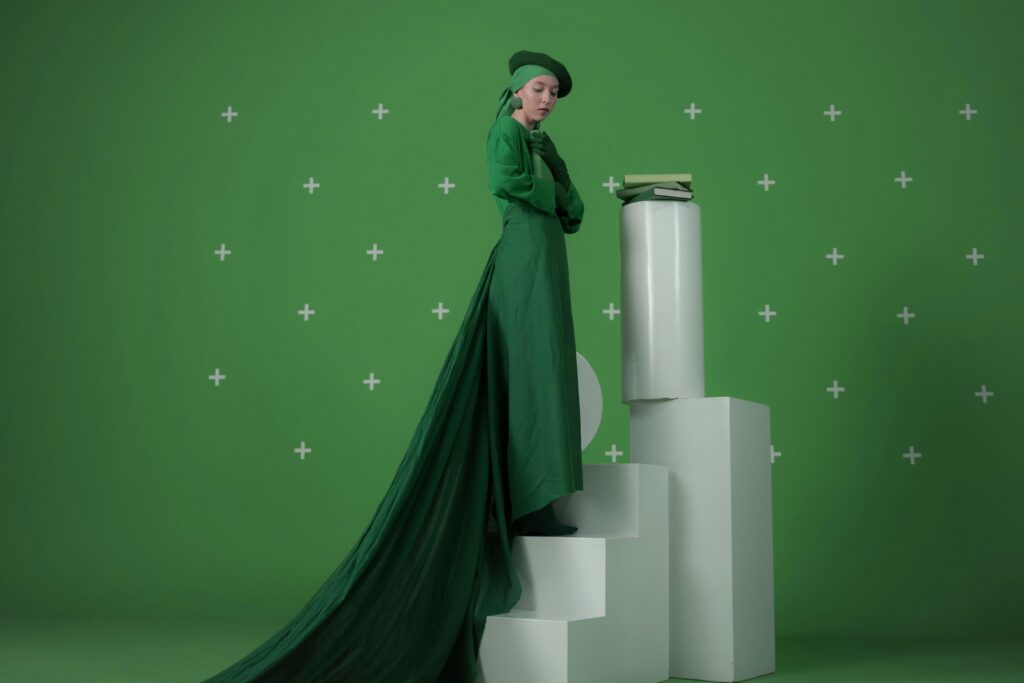 Elegant woman in a green dress standing on steps with books, against a green background.